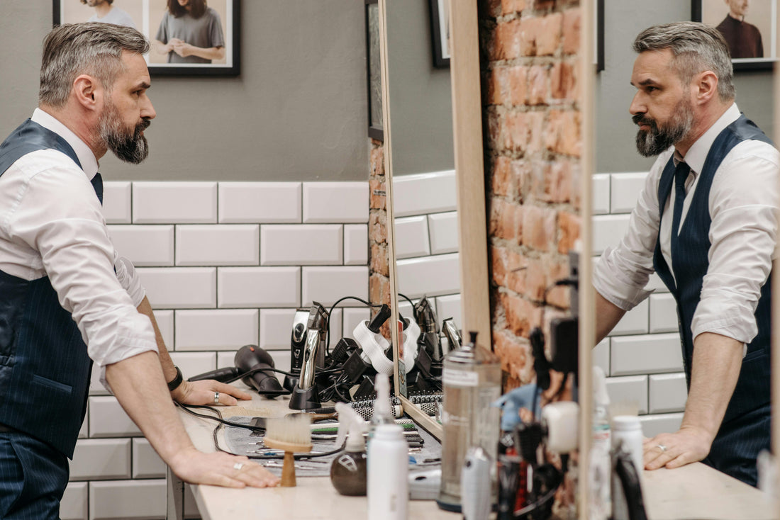Homme aux cheveux gris et barbe se regardant dans le miroir d’un salon de barbier, appuyé sur le comptoir entouré d’outils de coiffure.