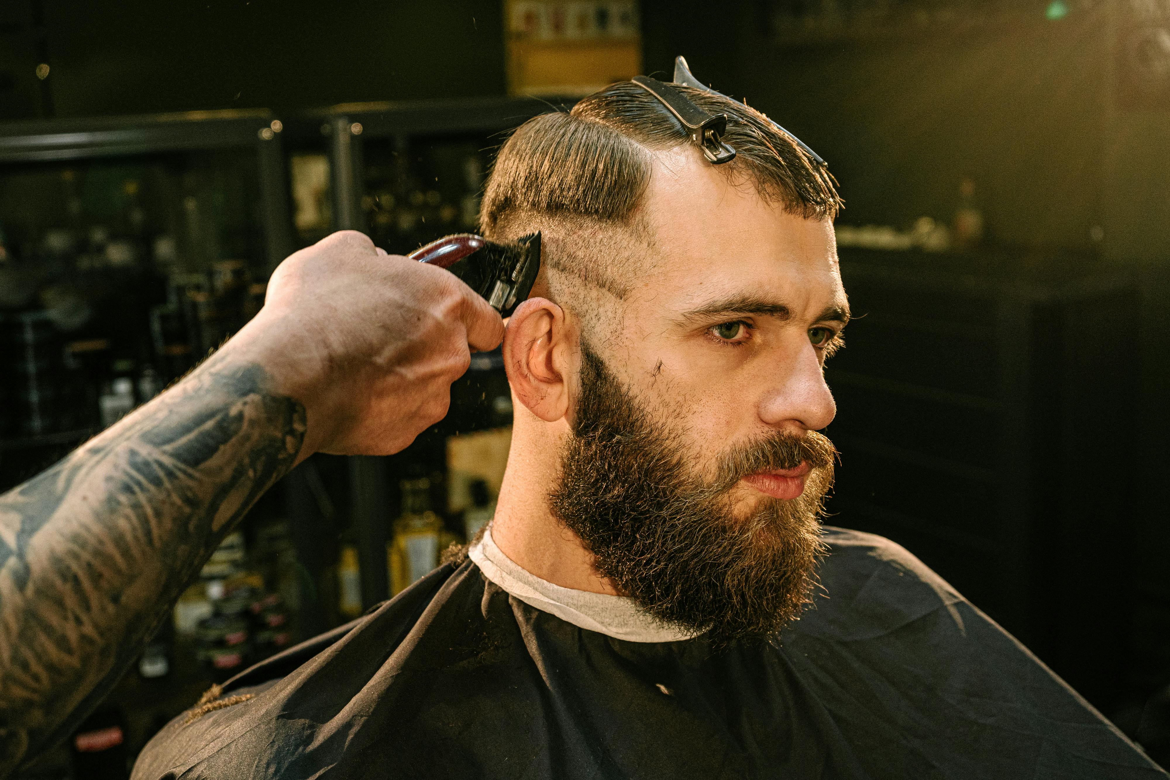 Homme se faisant couper les cheveux chez un barbier, utilisation de tondeuse pour réaliser un dégradé, avec une pince pour séparer les cheveux, gros plan sur la barbe et la coupe.